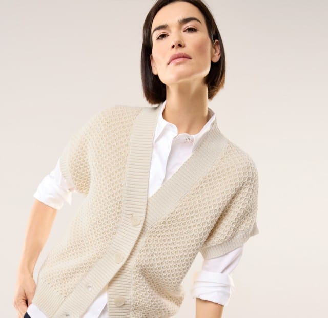 Eine Frau trägt eine Strickweste mit Waffelstruktur in Offwhite, darunter eine weiße Bluse.