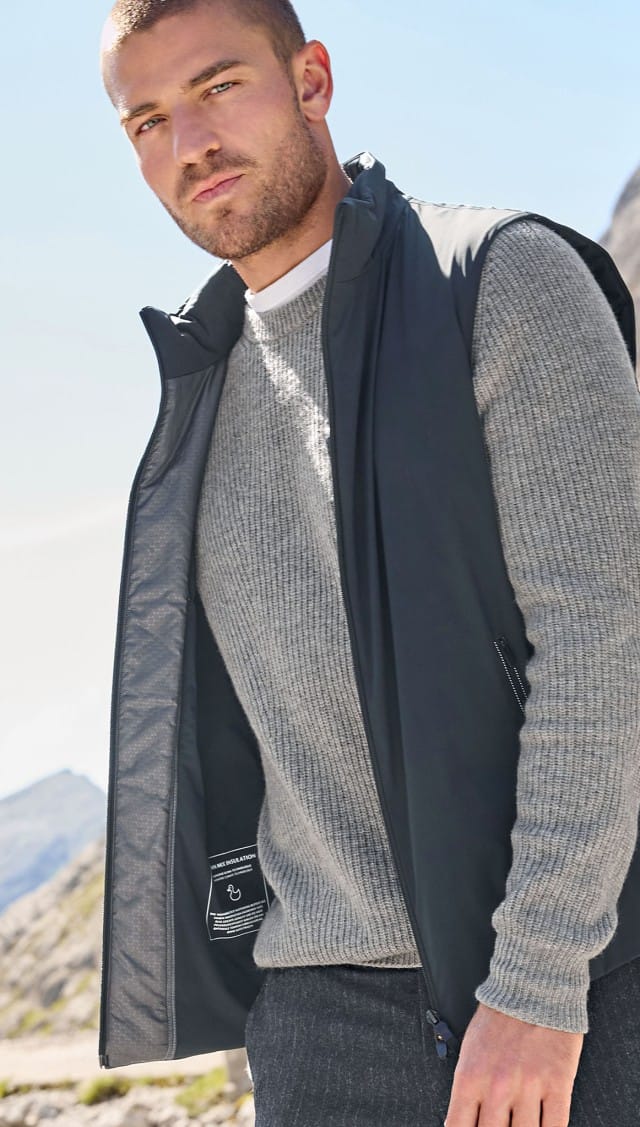 Ein Mann gesteht vor einem Gebirge und trägt eine Weste in Marine, darunter einen grauen Pullover.