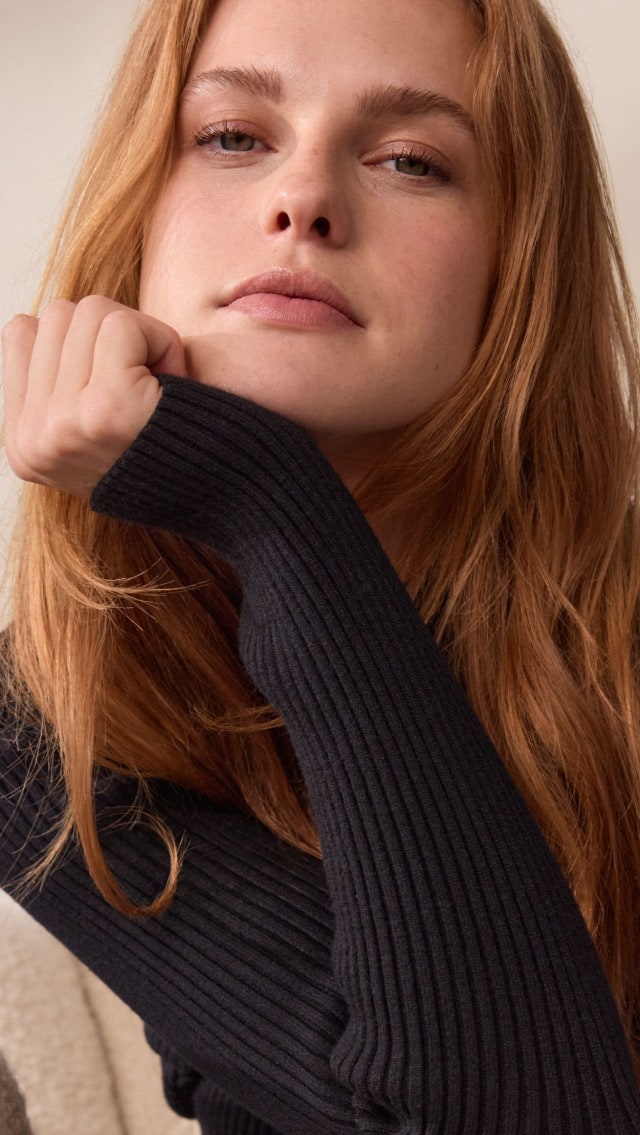Eine Frau trägt ein elegantes, schwarzes Strickkleid mit langen Ärmeln. Sie kombiniert den Look mit einer hellen Jacke oder warmen Stricksocken für einen gemütlich-modernen Stil.
