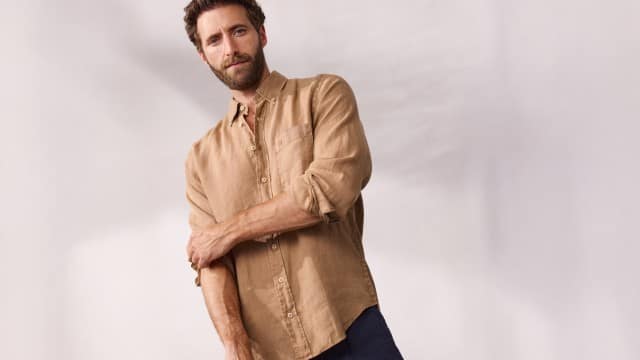 Eine Person steht vor einem hellen Hintergrund und trägt ein beiges Leinenhemd mit aufgekrempelten Ärmeln und eine dunkle Hose.