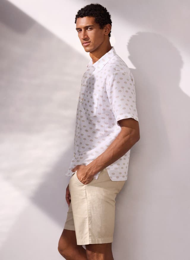 Ein Mann trägt ein weiß-bedrucktes Leinen-Mix Revers-Kragen-Hemd und hält die Hände in den Taschen einer Leinenmix Bermudas.