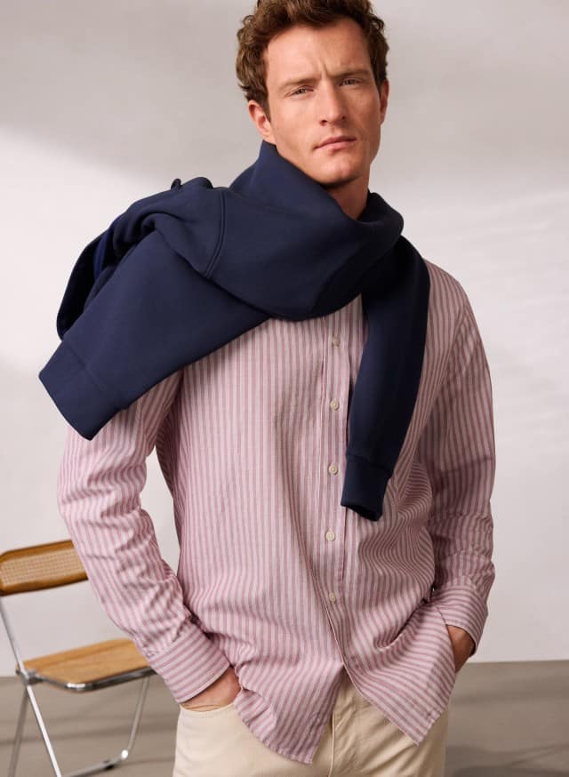 Ein Mann trägt Hemd aus Royal-Oxford mit roten Streifen und hat eine Hoodie quer über die Schultern gebunden.