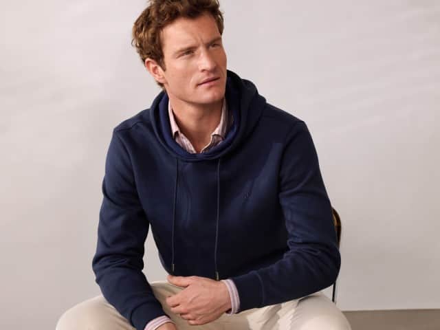 Ein Mann trägt einen Hoodie in Marine, darunter ein rot-gestreiftes Hemd und dazu eine helle Hose.