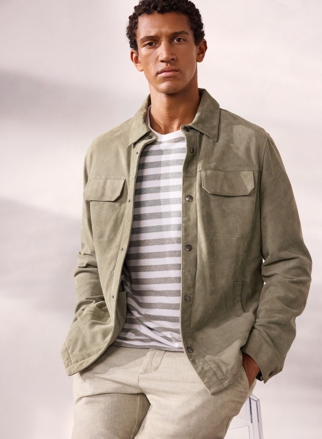 Ein Mann trägt eine Velourslederhemdjacke in der Farbe Salbei, darunter ein gestreiftes Shirt und dazu eine helle Hose.