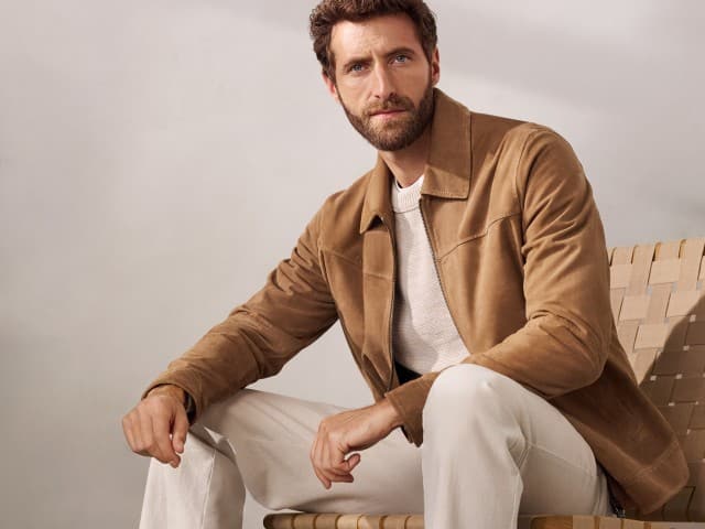 Ein Mann sitzt auf einem Sessel und trägt einen Ziegenvelours Blouson in Beige, darunter einen hellen Pullover und dazu eine helle Hose.