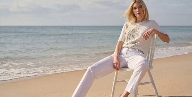 Frau in heller Sommerkleidung mit weißer Hose und T-Shirt mit Palmenmotiv sitzt lässig auf einem Stuhl am Strand. Der Stil wirkt entspannt und elegant.
