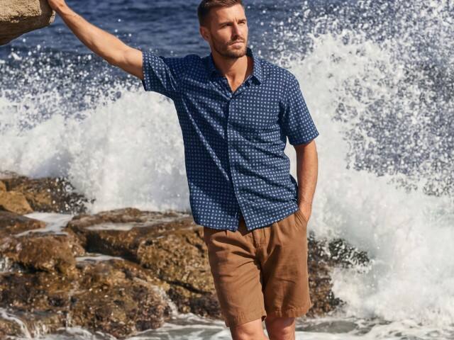 Mann in kurzärmeliger, dunkelblauer Hemdbluse mit dezentem Muster und brauner Shorts. Der Stil wirkt lässig, maritim und sommerlich.