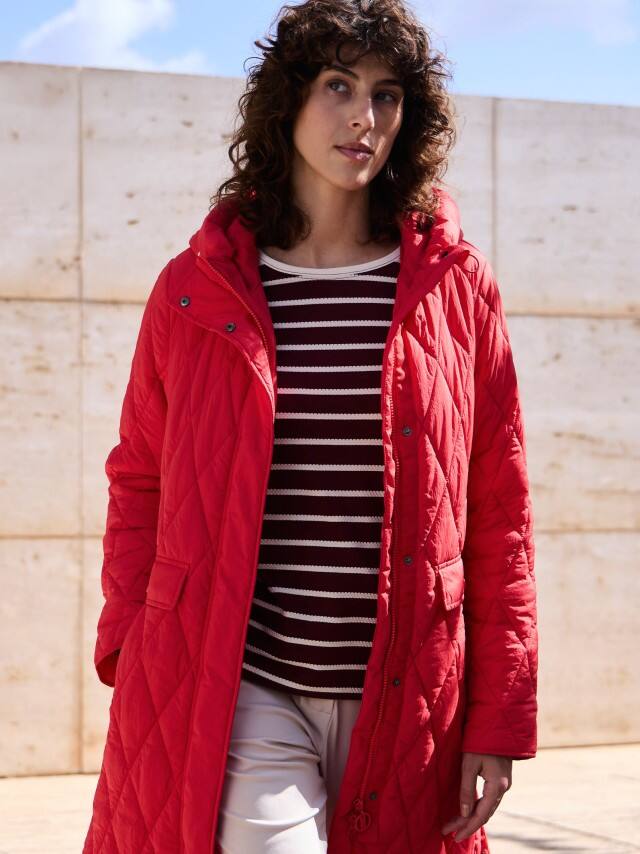 Eine Frau steht vor einer Natursteinwand und trägt einen Sandwich-Leicht-Steppmantel in Signalrot, darunter ein gestreiftes Baumwollshirt und dazu eine Jerseyhose.