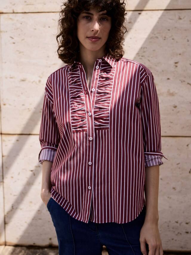 Ein Frau vor einer Natursteinwand trägt eine gestreifte Hemdbluse in Chianti.