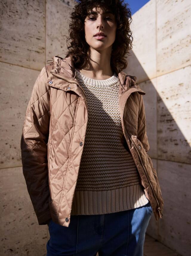 Eine Frau steht vor einer Natursteinwand und trägt eine Stepp Kurzjacke in der Farbe Praline, darunter einen passenden Pullover mit dezentem Streifen und dazu eine Marlene-Jeans.