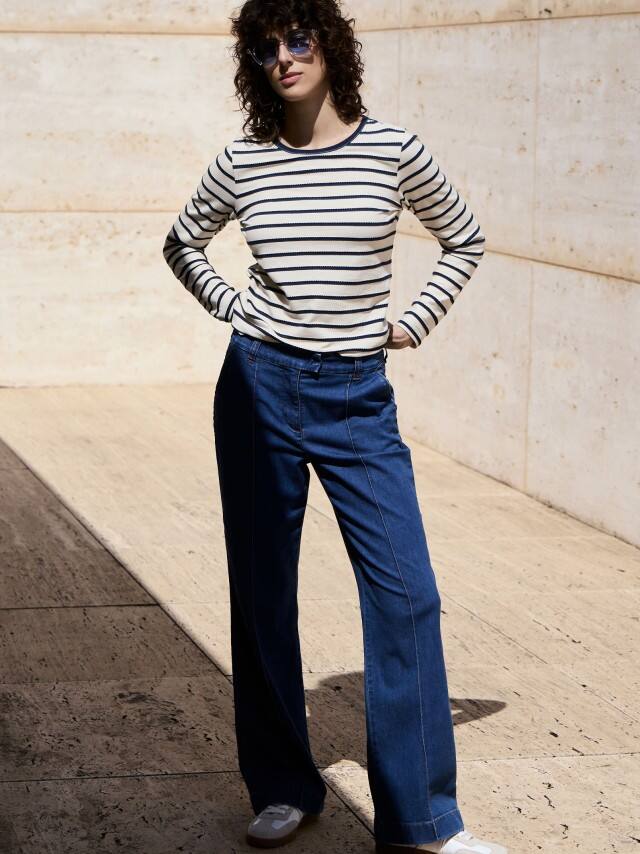 Eine Frau steht vor einer Natursteinwand und trägt ein Baumwollshirt Ringelstruktur in Weiß/Blau, dazu eine Marlene Jeans.