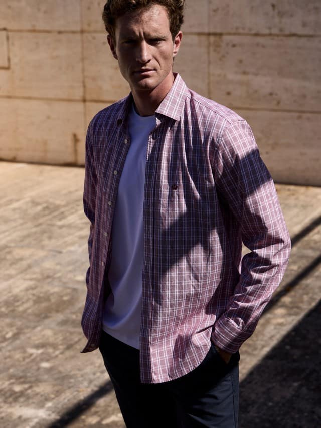 Ein Mann vor einer Natursteinwand trägt das Royal-Oxford Hemd in Karo-Rot, darunter ein weißes Shirt und dazu eine Chino in Beige.