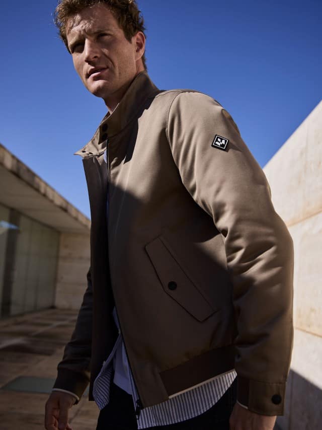 Ein Mann vor einem Natursteingebäude trägt einen graubraunen Freizeitblouson, dazu eine Jeans.