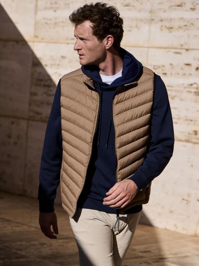 Ein Mann vor einer Natursteinwand trägt einen Hoodie in Marine, darüber eine Klima Leichtsteppweste in der Farbe Karamel, dazu eine helle Hose.