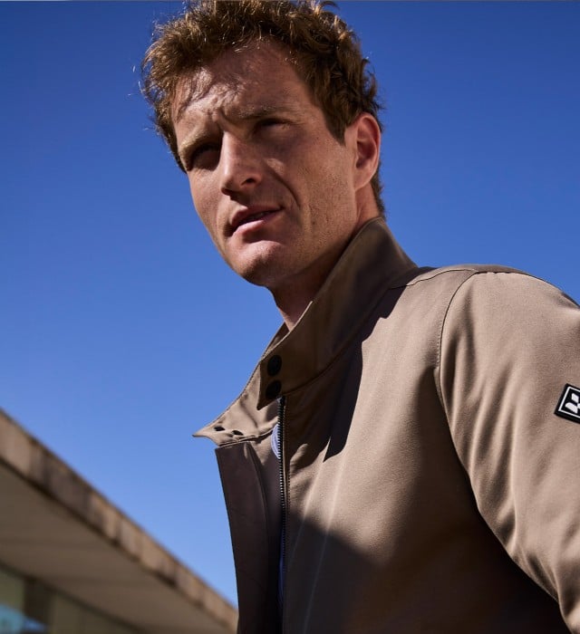 Ein Mann vor einem Natursteingebäude trägt einen graubraunen Freizeitblouson, dazu eine Jeans.