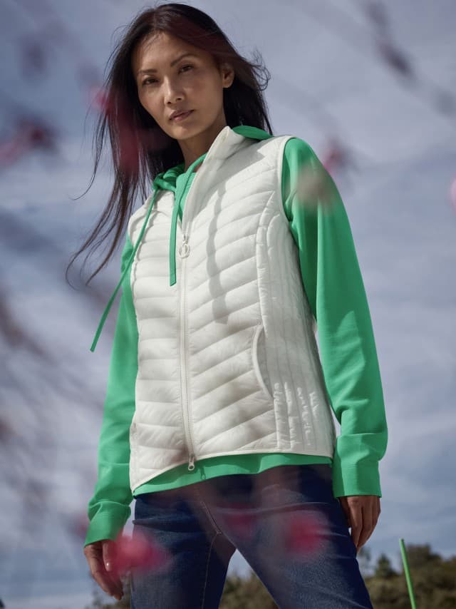 Eine Frau steht im Freien und trägt einen grünen Hoodie unter einer weißen Steppweste sowie eine Jeans. Eine Frau steht im Freien und trägt einen grünen Hoodie unter einer weißen Steppweste sowie eine Jeans.