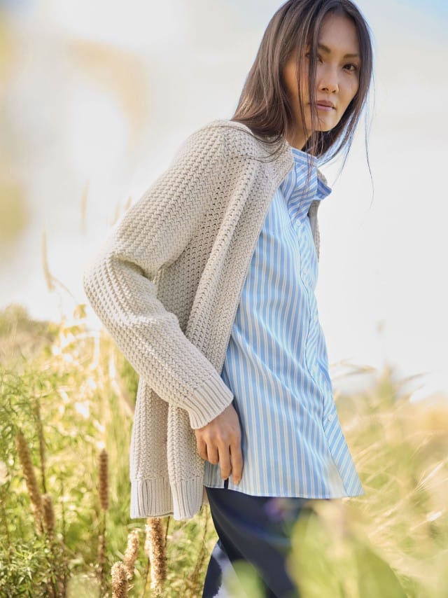 Eine Frau steht im Freien und trägt eine beige Strickjacke über einer hellblau gestreiften Bluse. Eine Frau steht im Freien und trägt eine beige Strickjacke über einer hellblau gestreiften Bluse.