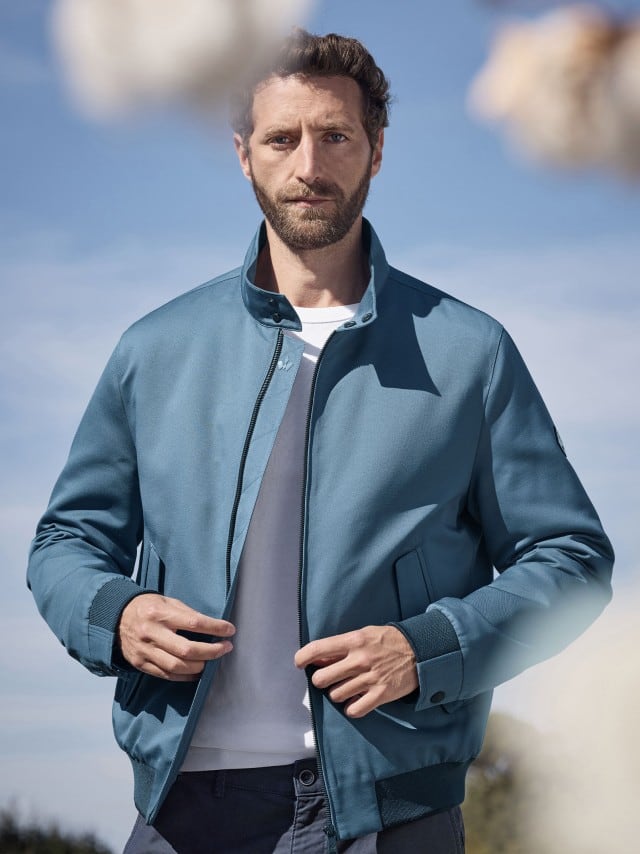 Ein Mann trägt einen Freizeitblouson in Rauchblau, darunter ein weißes Shirt. Im Vordergrund sind Blüten zu sehen. 