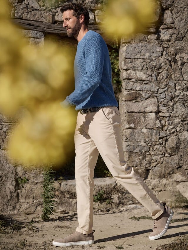 Ein Mann vor einer Steinwand trägt einen Strukturpullover Bicolor in Jeansblau, dazu eine helle Chino.