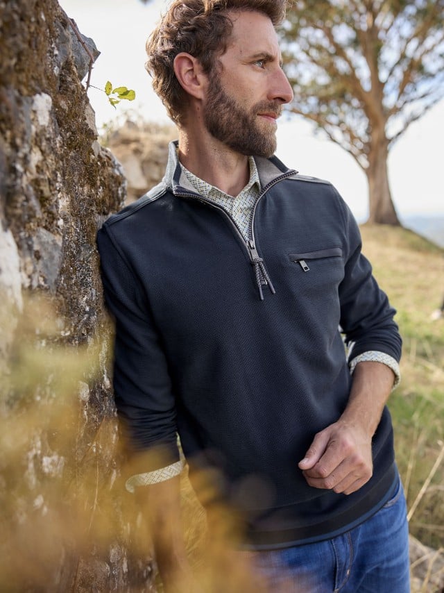 Ein Mann steht an einem Baum und trägt den Easycare Struktur-Troyer Bicolor in Marine, darunter ein Hemd und dazu eine Jeans.