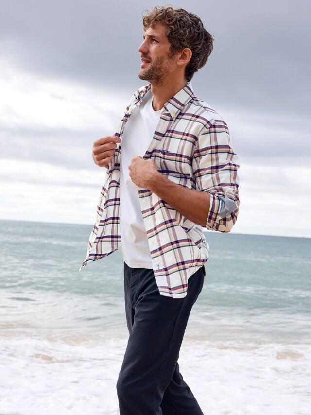 Ein Mann am Strand trägt ein kariertes Hemd in Offwhite, darunter ein weißes Shirt und dazu eine Chino in Marine.