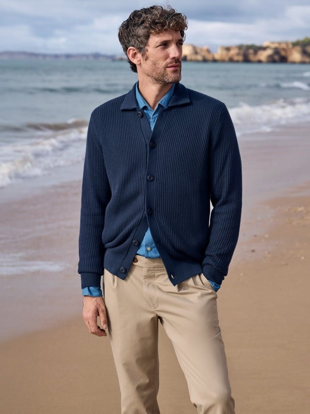 Ein Mann steht am Strand und trägt einen gerippten Polo-Cardigan in Marine, darunter eine Jeans-Hemd und dazu eine Chino in Beige.