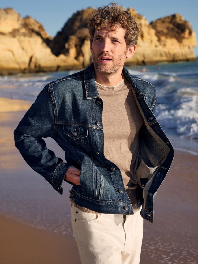 Ein Mann am Strand trägt eine Jeansjacke in Blau, darunter einen Premium Pullover und dazu eine weiße Charakter-Jeans.