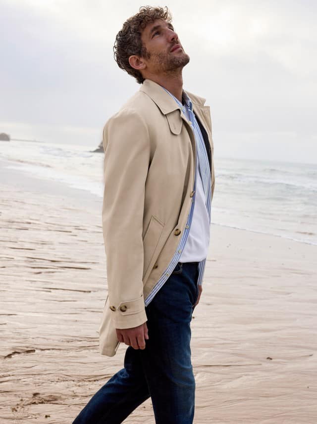 Ein Mann am Strand trägt einen Baumwoll Mantel in Beige, darunter ein Hemd und dazu eine Charakter-Jeans.