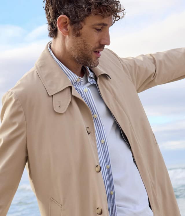 Ein Mann trägt einen beigen Mantel über einem gestreiften Hemd und einem weißen T-Shirt. Der Stil wirkt klassisch und lässig, ideal für einen Spaziergang am Meer.