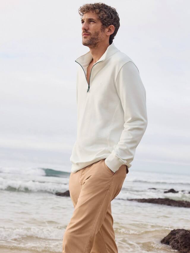 Ein Mann steht am Strand und trägt die 100 Komplimente Chino in Sandgelb, dazu einen Essential Troyer in Offwhite.
