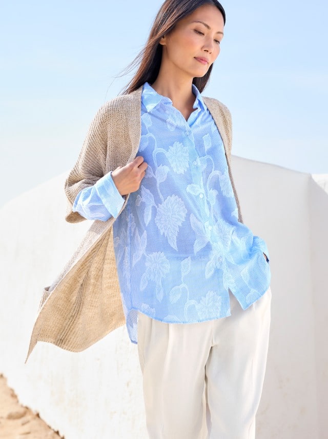 Eine Frau trägt eine locker fallende, hellblaue Bluse mit floralen Mustern zu heller Hose und Strickjacke, was leicht, frisch und entspannt wirkt. Eine Frau trägt eine locker fallende, hellblaue Bluse mit floralen Mustern zu heller Hose und Strickjacke, was leicht, frisch und entspannt wirkt.
