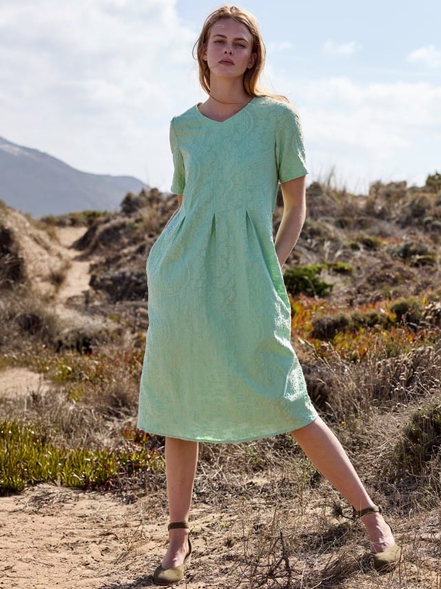 Eine Frau in einer felsigen Landschaft, trägt ein grünes Jacquardkleid und Sandalen mit Keilabsatz.