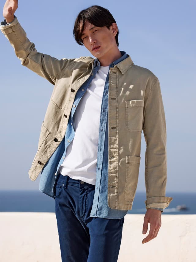 Ein Mann trägt eine beige Jacke über einem Jeanshemd und weißem T‑Shirt zu dunkelblauen Hosen, was lässig, modern und sommerlich entspannt wirkt.