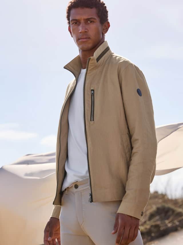 Ein Mann steht in einer Düne und trägt einen Leinenblouson, darunter ein weißes Shirt und dazu eine Baumwoll Five-Pocket in Safari-Beige.