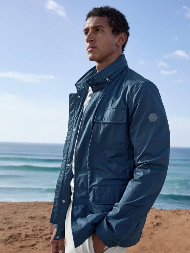 Ein Mann steht am Strand und trägt die Multitaschen Jacke in Marine, darunter ein gestreiftes Shirt und dazu ein weiße Hose.