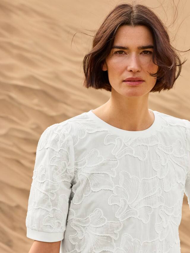 Eine Frau steht in den Dünen und trägt ein weißen Shirt mit Stickerei.
