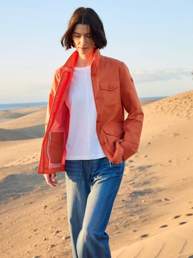 Eine Frau steht in den Dünen und trägt einen Jacke in Terra, darunter ein weißes Shirt und dazu eine Jeans.