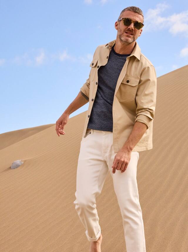 Ein Mann steht in den Dünen und trägt eine helle Hemdjacke, darunter ein Shirt und dazu eine weiße Hose.
