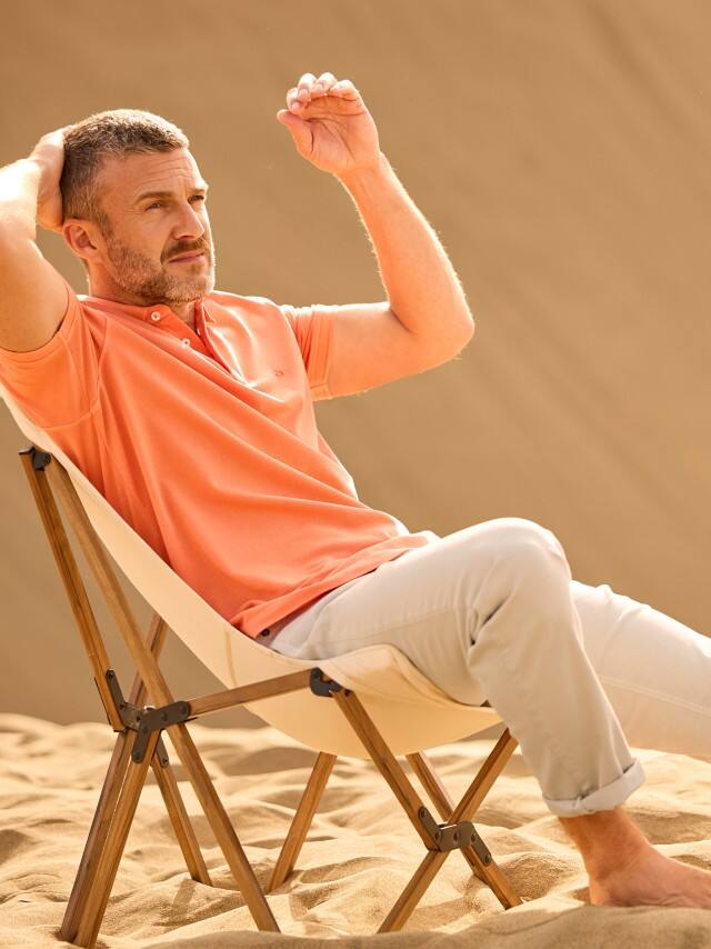 Ein Mann trägt ein Poloshirt in Orange, dazu eine helle Hose und sitzt auf einem Stuhl in den Dünen.