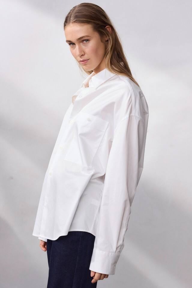 Eine Frau trägt ein weißes Hemd und steht vor einem neutralen Hintergrund.