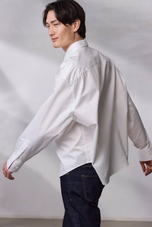 Ein Mann trägt ein weißes Hemd und dunkle Jeans und geht vor einem neutralen Hintergrund.