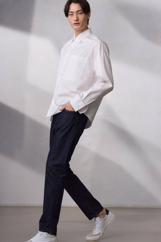 Ein Mann geht vor einem hellen Hintergrund und trägt ein weißes Hemd, dunkle Jeans und weiße Sneaker.