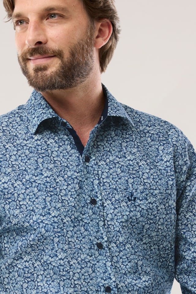 Ein Mann trägt ein blaues Hemd mit dichtem floralem Muster und blickt leicht zur Seite.
