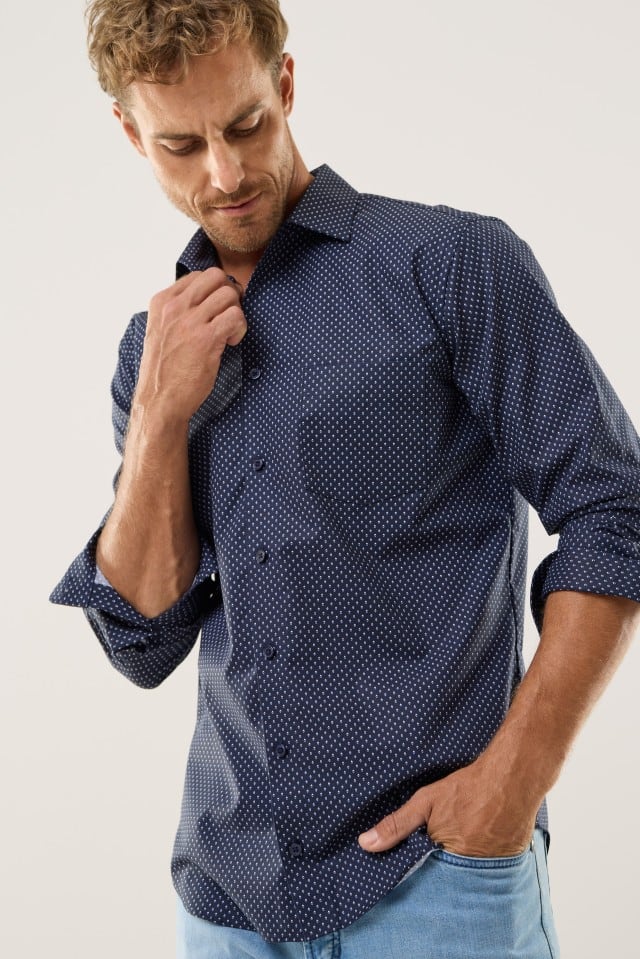 Auf dem Bild ist eine Person mit kurzen, hellbraunen Haaren zu sehen, die ein dunkelblaues Hemd mit feinem weißen Punktmuster und eine hellblaue Jeans trägt.