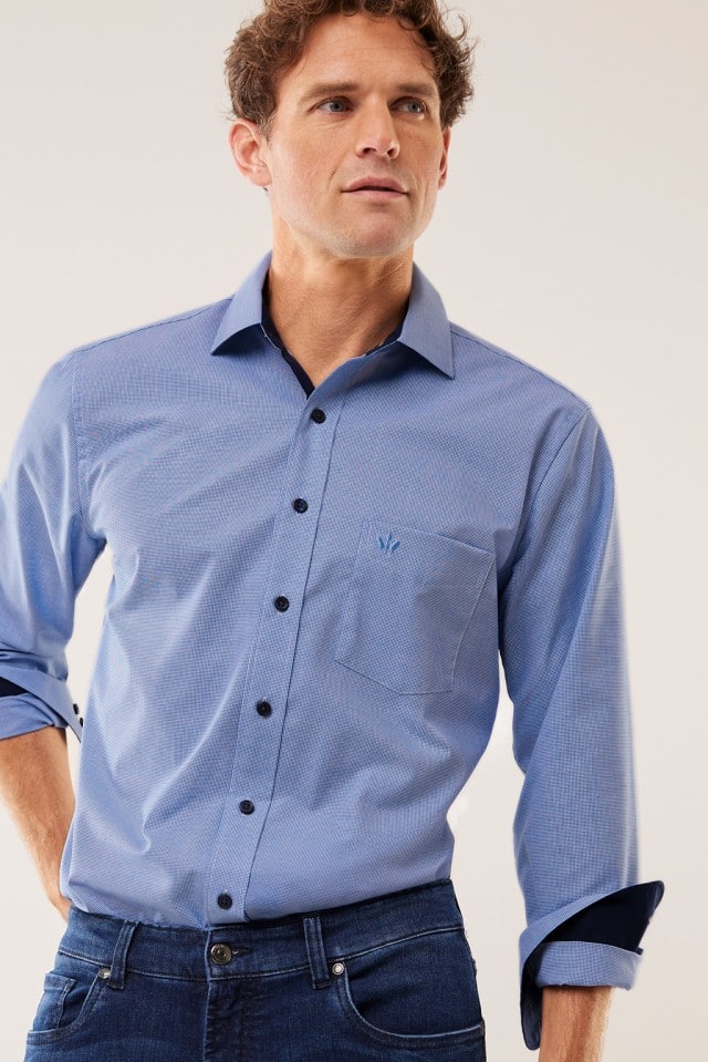 Ein Mann trägt ein blaues langärmliges Hemd und eine dunkle Jeans vor neutralem Hintergrund.