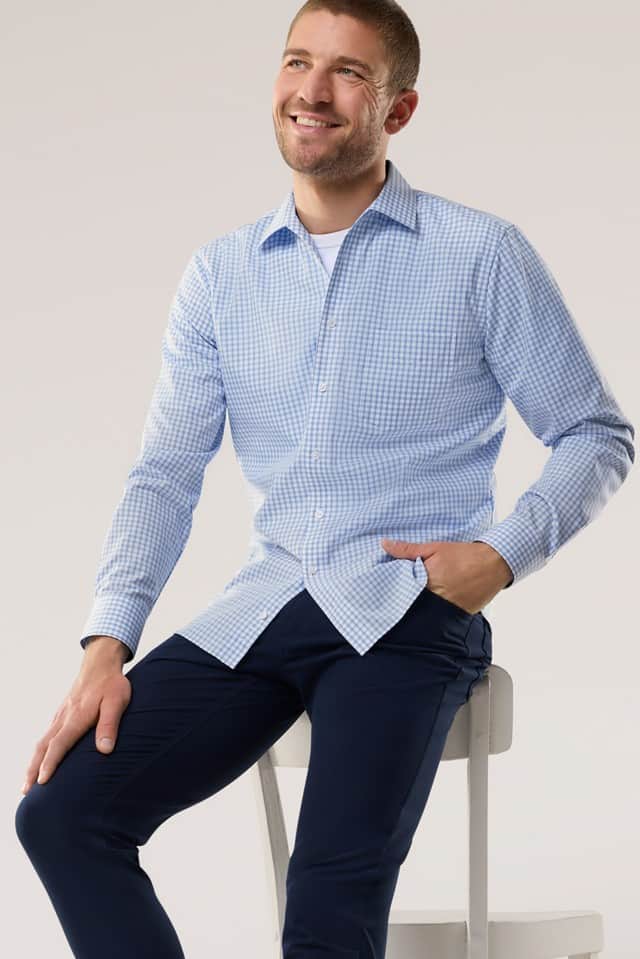 Eine Person sitzt entspannt auf einem Hocker und trägt ein hellblau kariertes Hemd sowie eine dunkelblaue Hose.