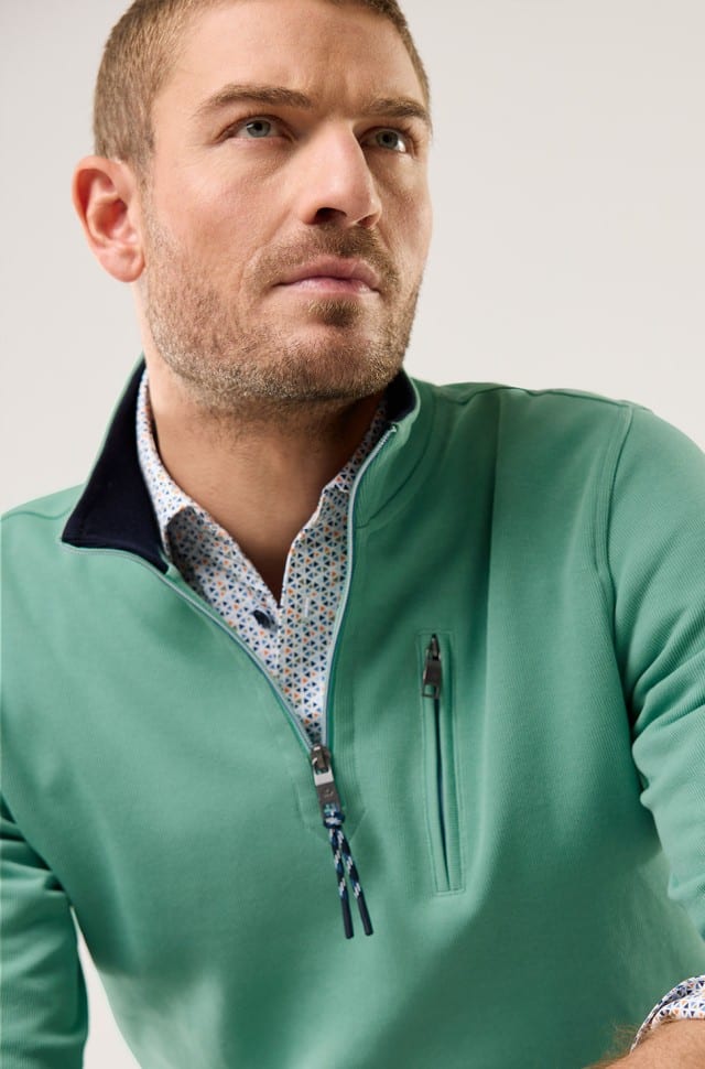Ein Mann trägt einen grünen Pullover mit kurzem Reißverschluss über einem gemusterten Hemd und blickt zur Seite.