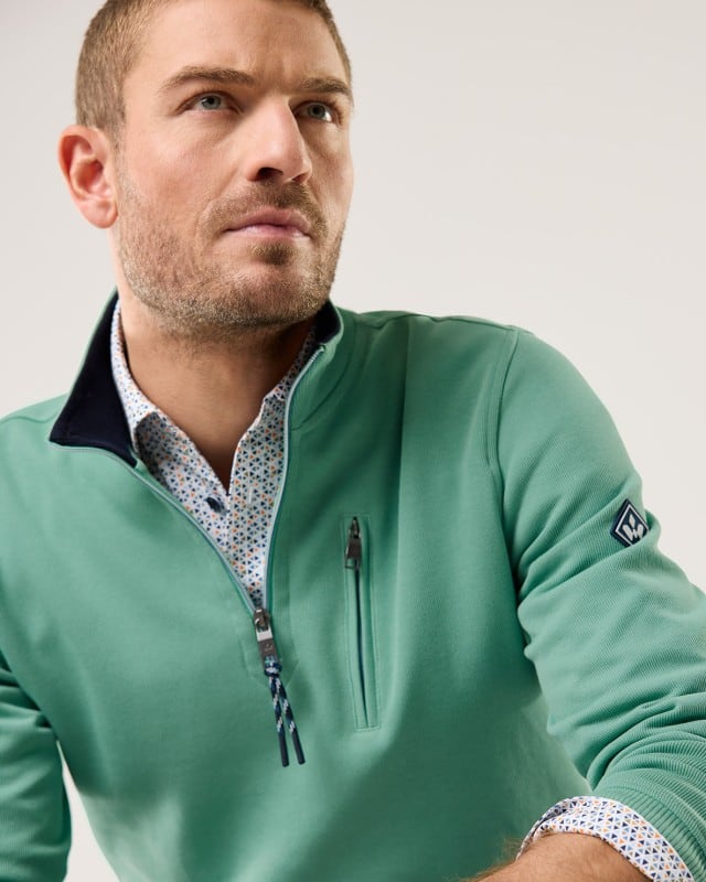 Ein Mann trägt einen grünen Pullover mit kurzem Reißverschluss über einem gemusterten Hemd und blickt zur Seite. Ein Mann trägt einen grünen Pullover mit kurzem Reißverschluss über einem gemusterten Hemd und blickt zur Seite.