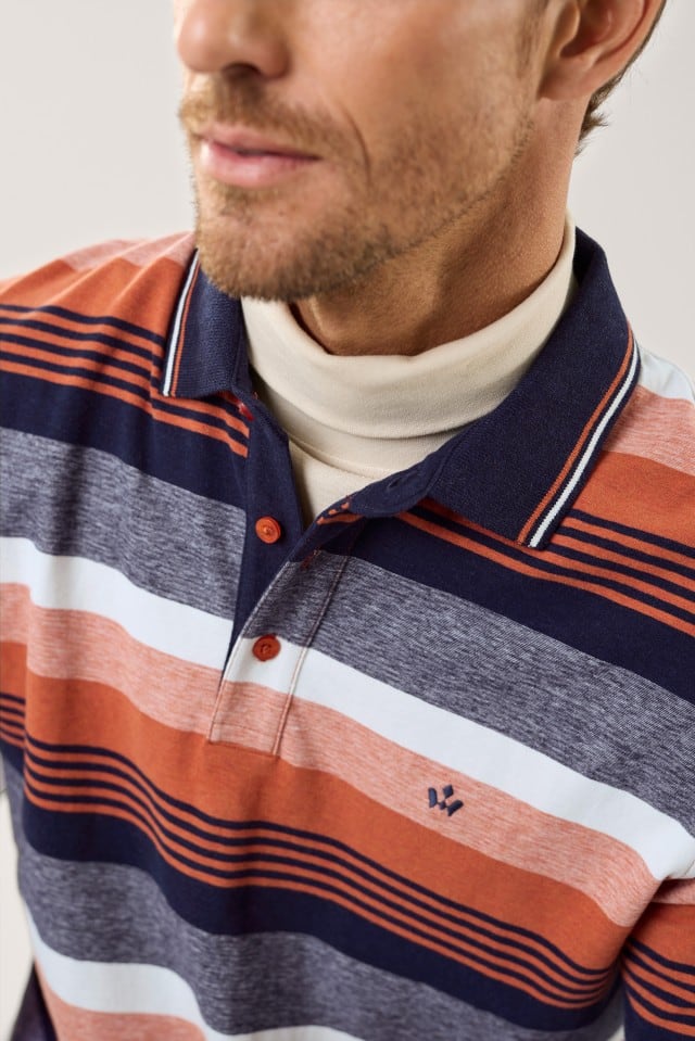 Ein Mann trägt ein langärmliges Poloshirt mit breiten Querstreifen in Orange-, Blau-, Grau- und Weißtönen; der Kragen hat feine Streifendetails, und darunter ist ein cremefarbener Rollkragenpullover sichtbar.