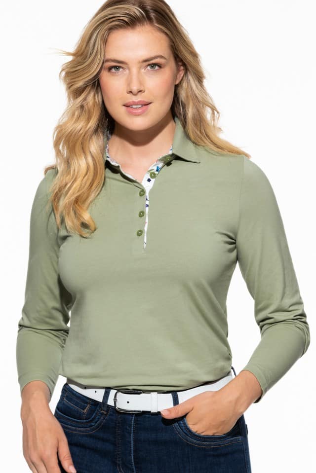 Eine Frau mit langen, blonden Haaren trägt ein langärmliges, hellgrünes Poloshirt mit gemustertem Stoffeinsatz an der Knopfleiste und kombiniert es mit einer dunklen Jeans und einem weißen Gürtel.